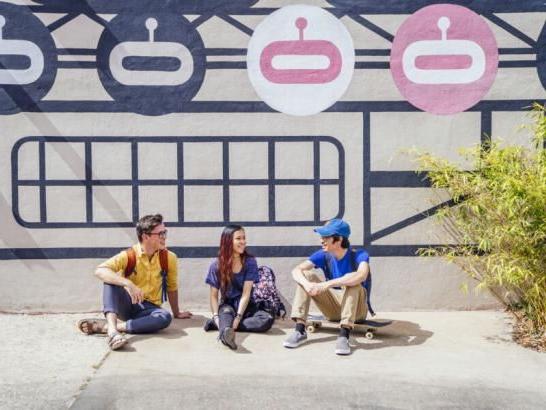 Three students sit on a sidewalk on South Congress Avenue in front of a black, white and pink mural.